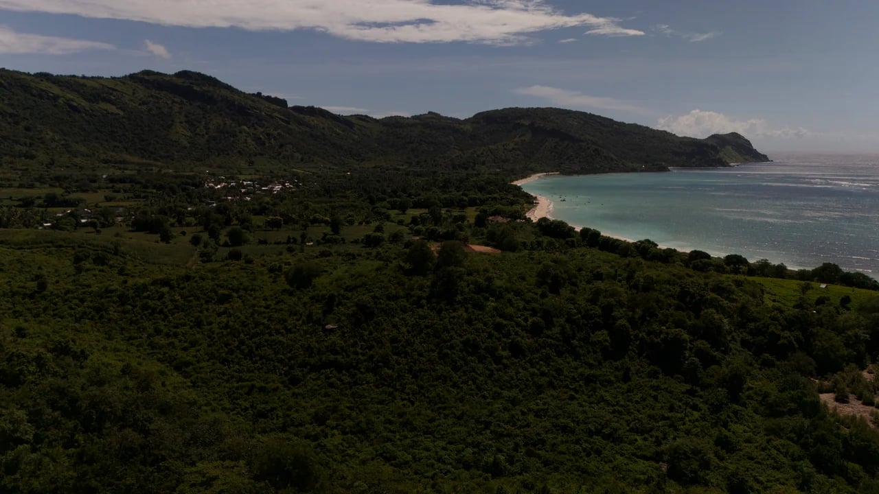 Wide coastal drone shot of the curved bay, sandy beach, lush green hills and distant mountains of West Sumbawa